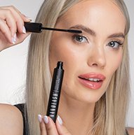 Uma mulher aplica rímel com cuidado nos cílios, segurando o pincel bem próximo para um acabamento perfeito.